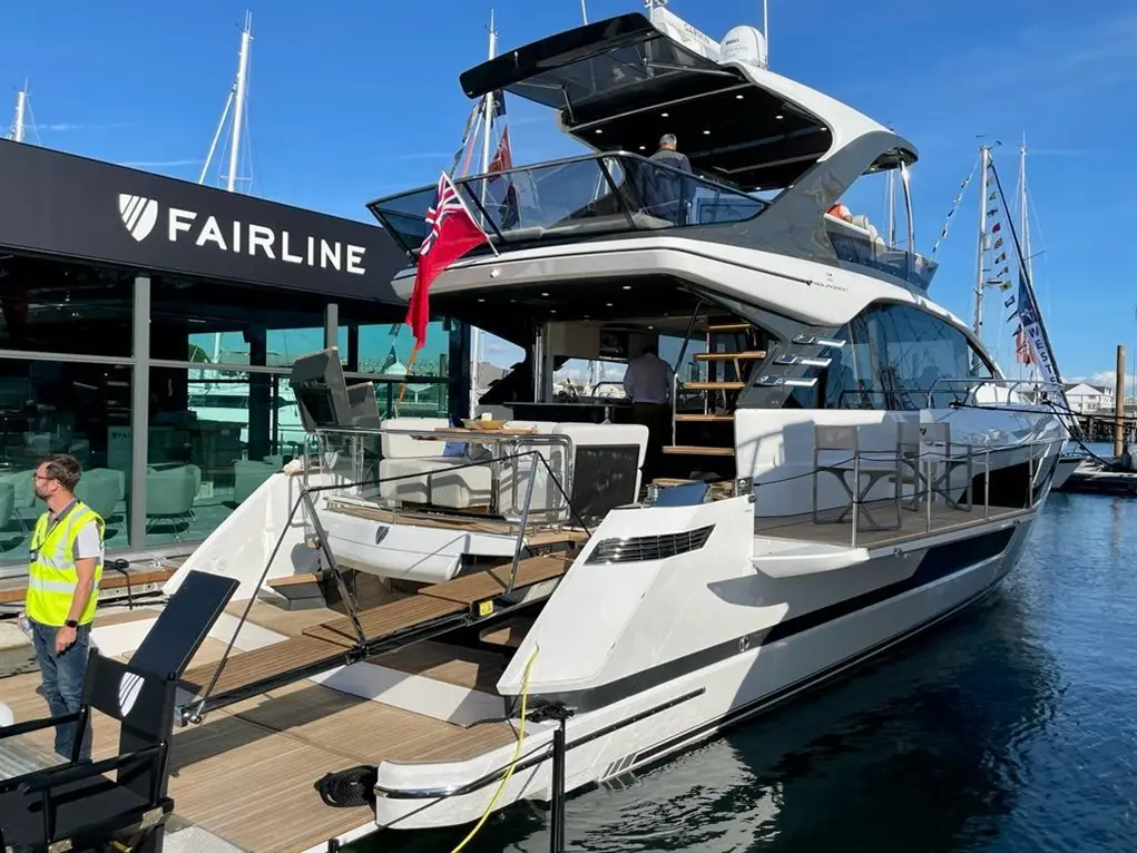 A luxury Fairline yacht docked at a marina, featuring a seating area, stairs, and a man in a high-visibility vest nearby.